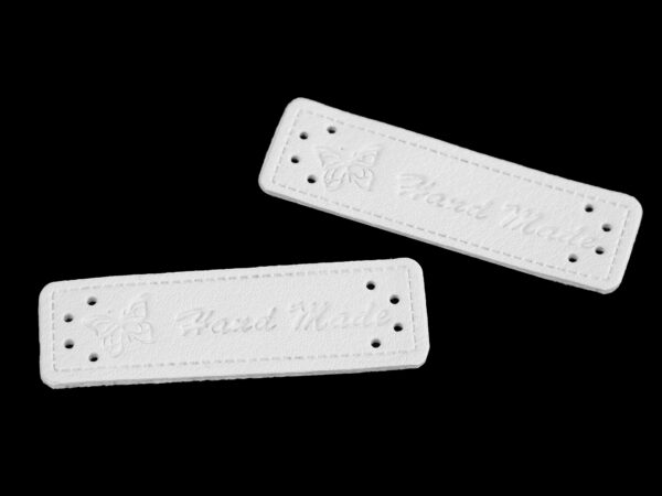 Valkoinen Hand made -merkki, jossa on vasemmassa reunassa koristeena perhonen. Mitat: 5 x 1,5 cm. Paksuus 1,2 mm. Molemmissa päissä on neljä reikää kiinniompelua varten.
