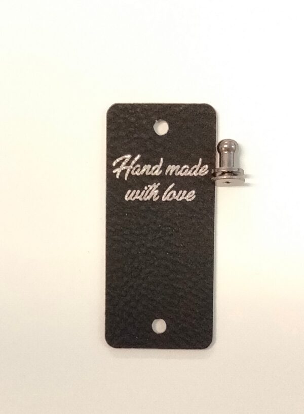 Merkki teksillä "Hand made with love". Musta, tekonahkaa. Merkin voi kiinnittää ja irrottaa ruuvattavalla nastalla. Koko: 2,5 cm x 5,8 cm